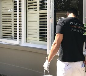 Painter applying white gloss to window frames set against freshly painted Dulux Gnu Tan exterior walls in Castle Cove