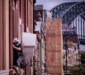 Rope access painter working on window frames at the Fortune of War Hotel in The Rocks, with Sydney Harbour Bridge visible.